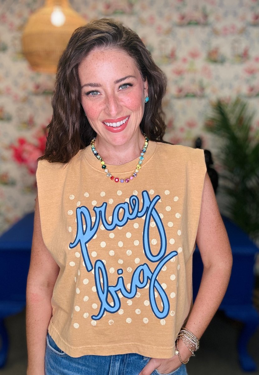 A smiling woman wearing a tan sleeveless crop top with blue script reading 'pray big' and white polka dots, paired with blue jeans, in a decorated indoor setting.