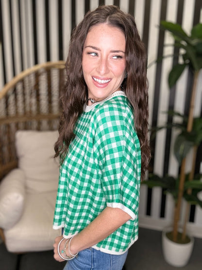 Woman wearing a green and white checkered shirt in a casual setting with a plant and chair.