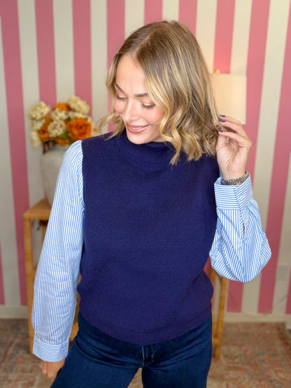 A woman wearing a navy blue ribbed turtleneck sweater vest over a light blue and white striped button-down shirt, paired with dark blue jeans, standing in front of a pink and white striped wall.