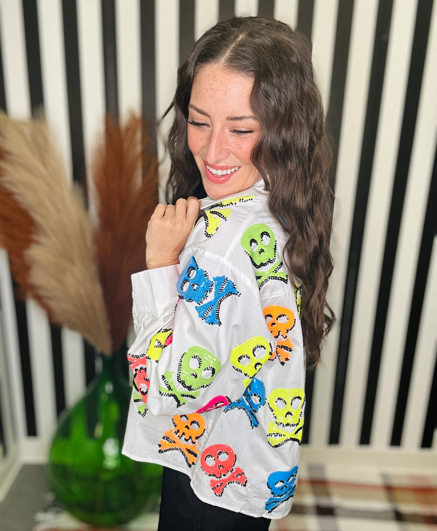 A woman wearing a white long-sleeve button-up shirt covered in colorful skull and crossbones sequin patches, posed in front of a black and white striped background.