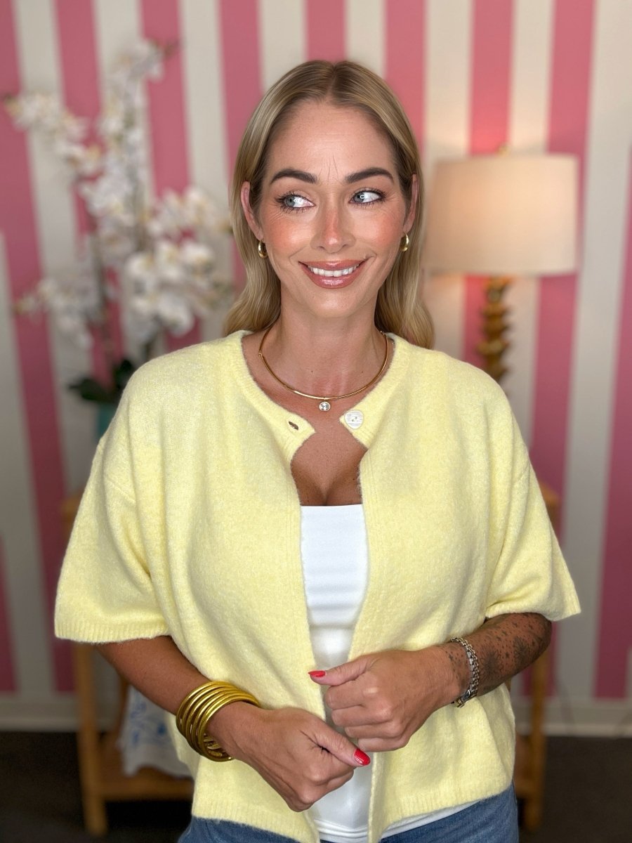 A woman wearing a pale yellow short-sleeve knit cardigan over a white top, standing in front of a pink and white striped wall with a lamp in the background.