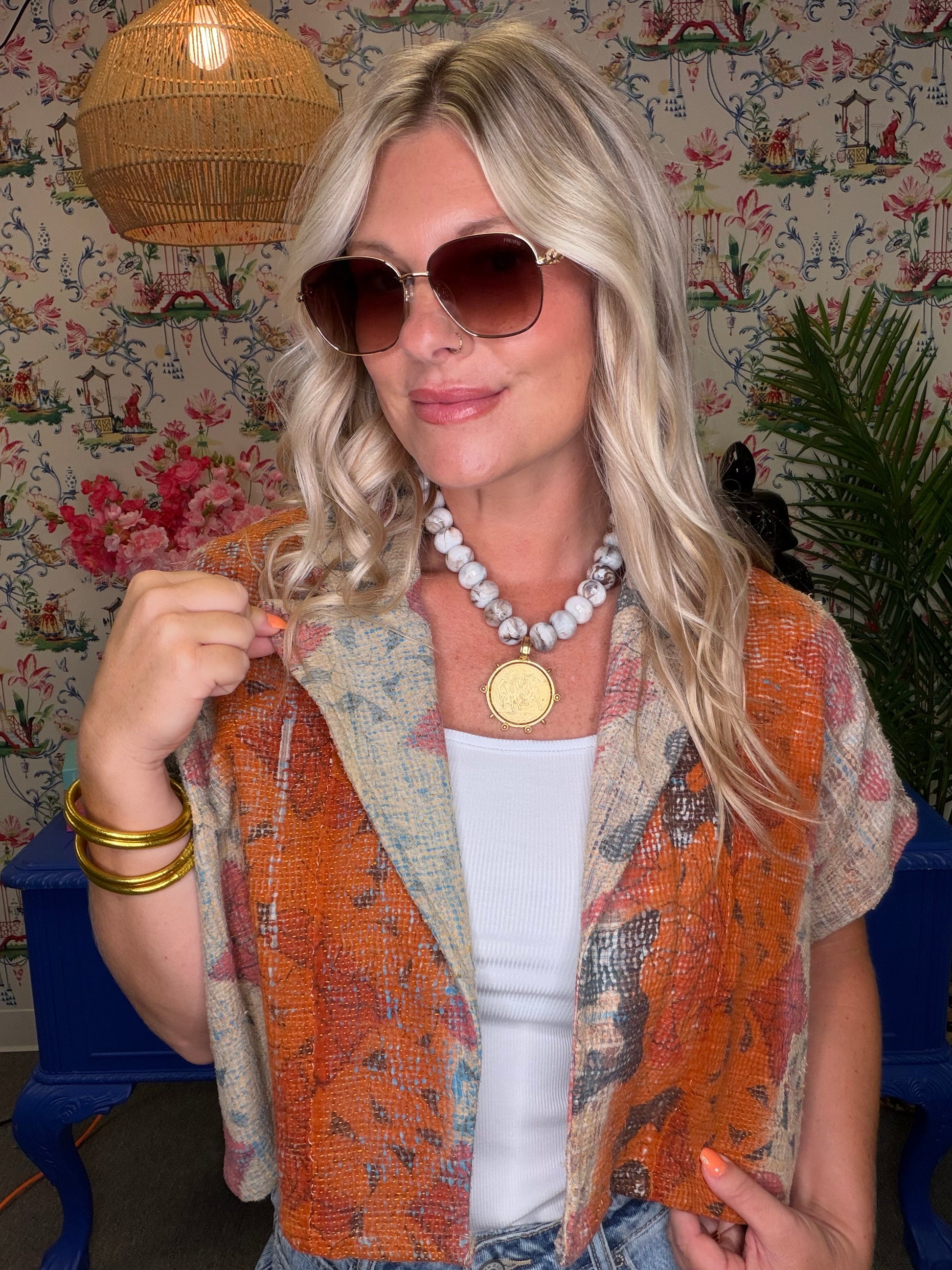 A woman with blonde hair wearing a short-sleeve orange and beige patterned vest over a white top, styled with sunglasses, a chunky white beaded necklace with a gold medallion, and gold bracelets, set against a floral wallpaper background.