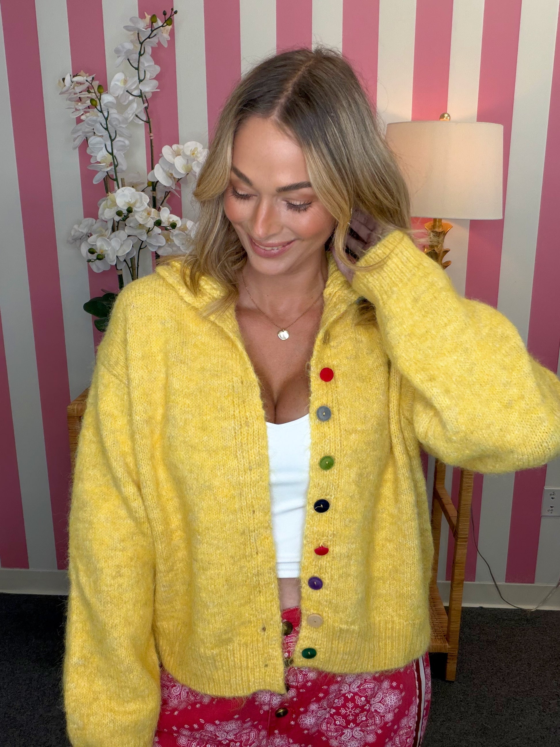 A woman wearing a fuzzy yellow knit cardigan with colorful buttons over a white top and pink floral pants, standing in front of a pink and white striped wall.