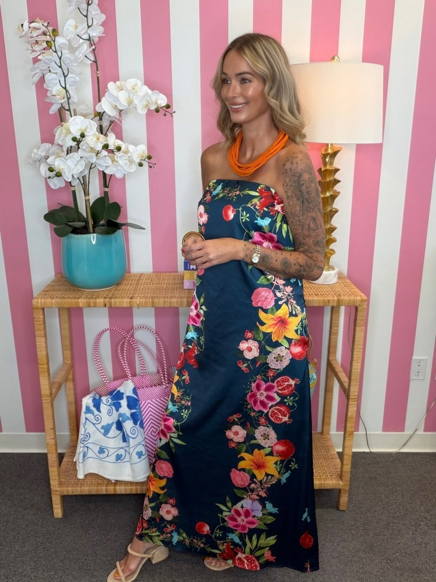 A woman modeling a strapless navy maxi dress with a vibrant multicolor floral print, standing in front of a pink and white striped wall next to a lamp and a side table.