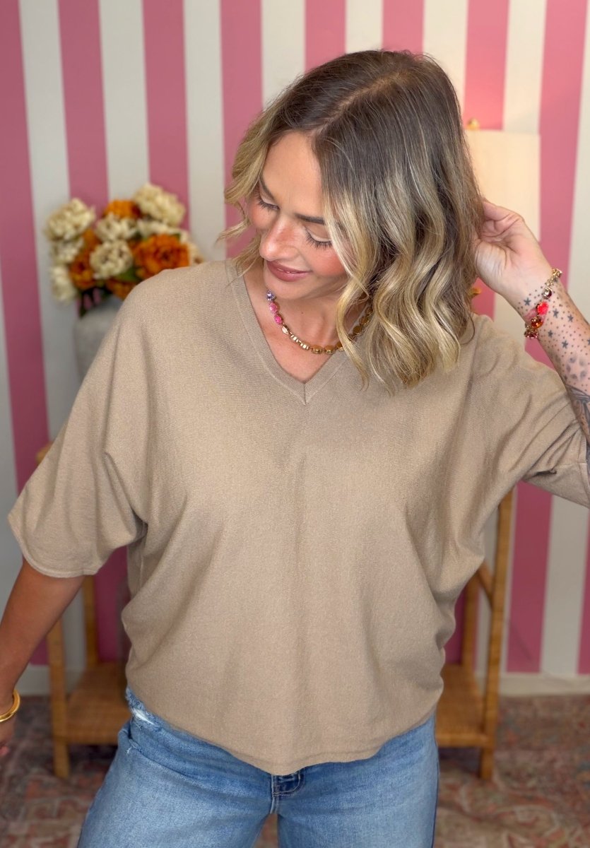 A woman wearing a beige short-sleeve V-neck knit top and blue jeans, standing in front of a pink and white striped wall.
