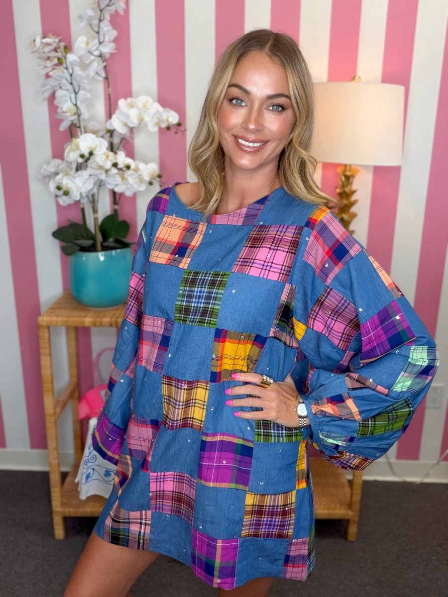 A woman modeling a blue patchwork plaid tunic dress in a pink and white striped room with a wooden side table and lamp.