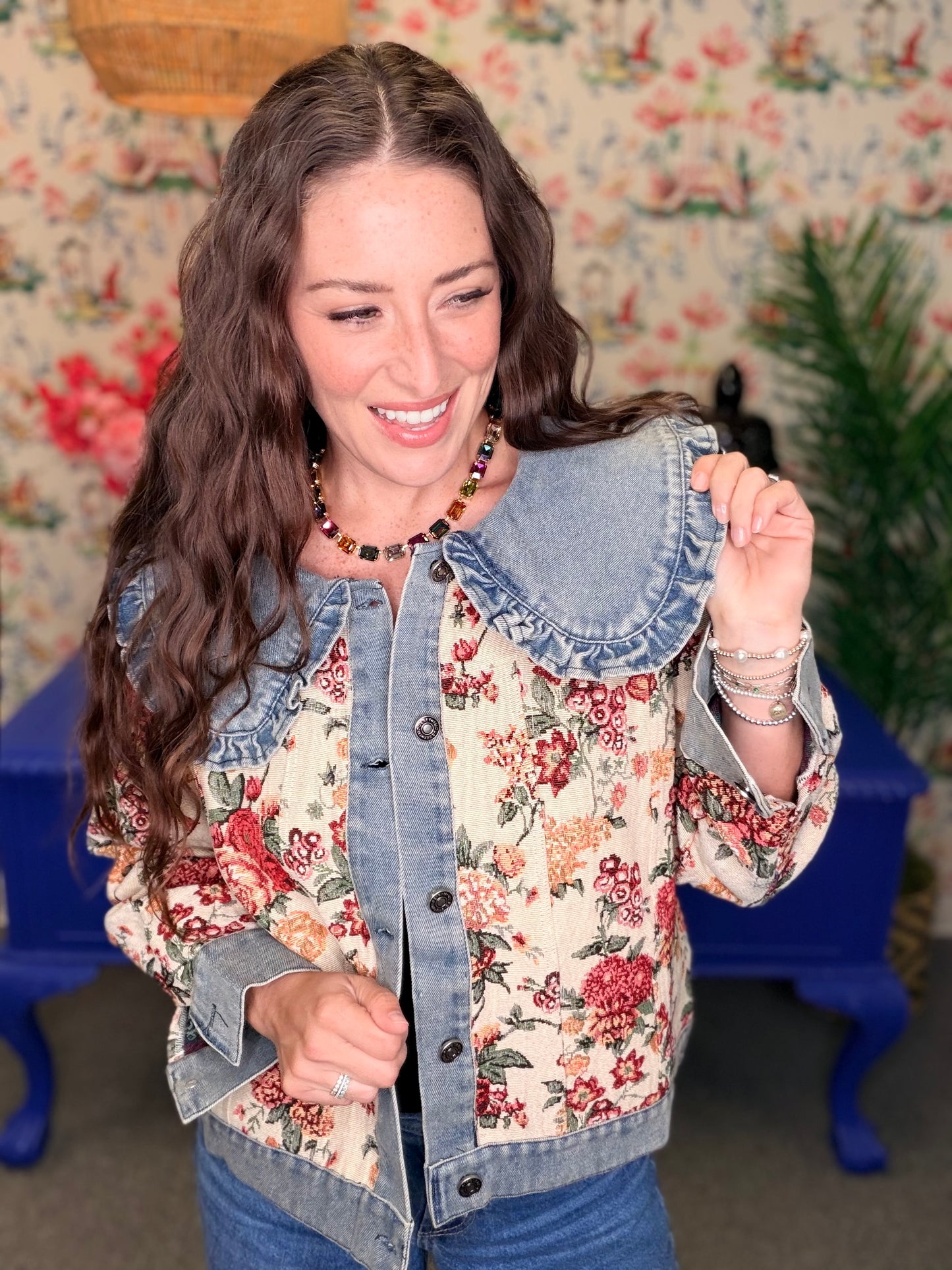 A woman smiling and wearing a hybrid denim jacket with a large denim collar and a red floral print body, posed in front of a vintage floral wallpaper background.
