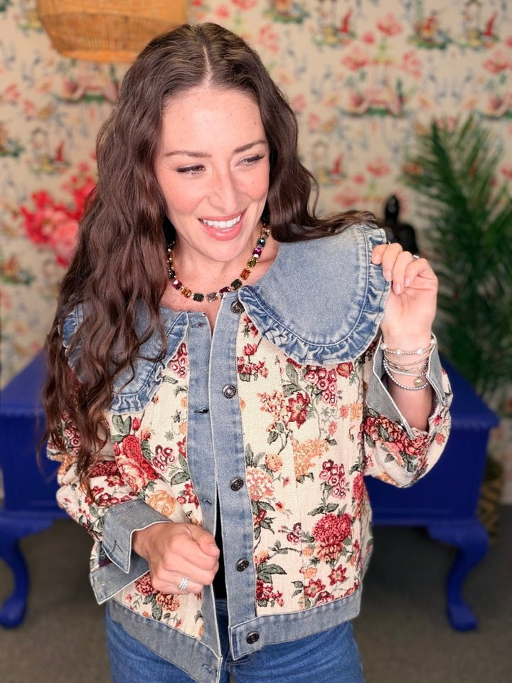 A woman smiling and wearing a hybrid denim jacket with a large denim collar and a red floral print body, posed in front of a vintage floral wallpaper background.