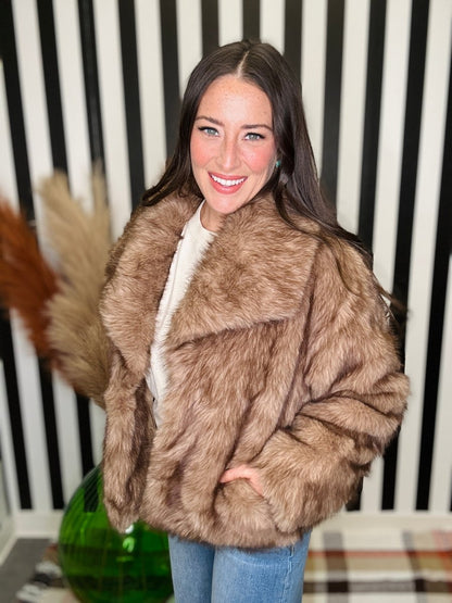 A woman wearing a voluminous brown faux fur coat over a white top and blue jeans, standing in front of a black and white striped wall.