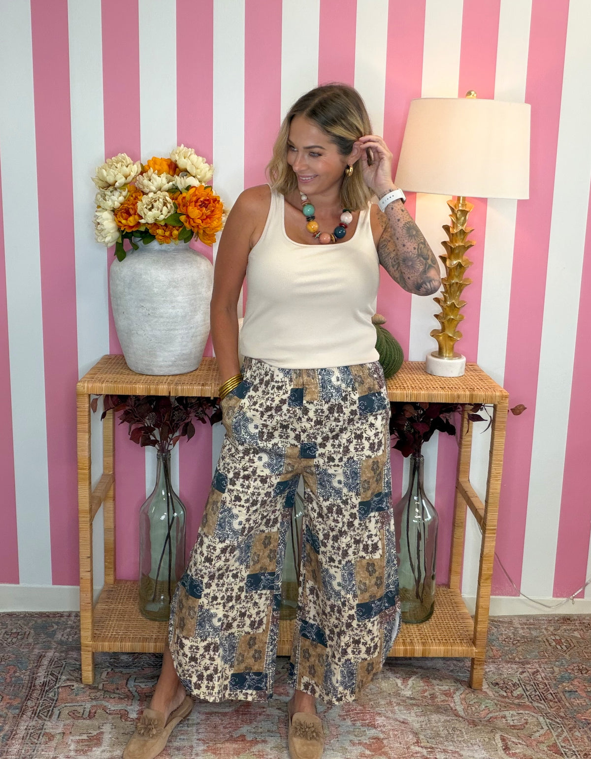 A woman modeling a cream ribbed tank top and beige wide-leg patchwork pants with blue, brown, and black animal print details, standing in front of a pink and white striped wall.