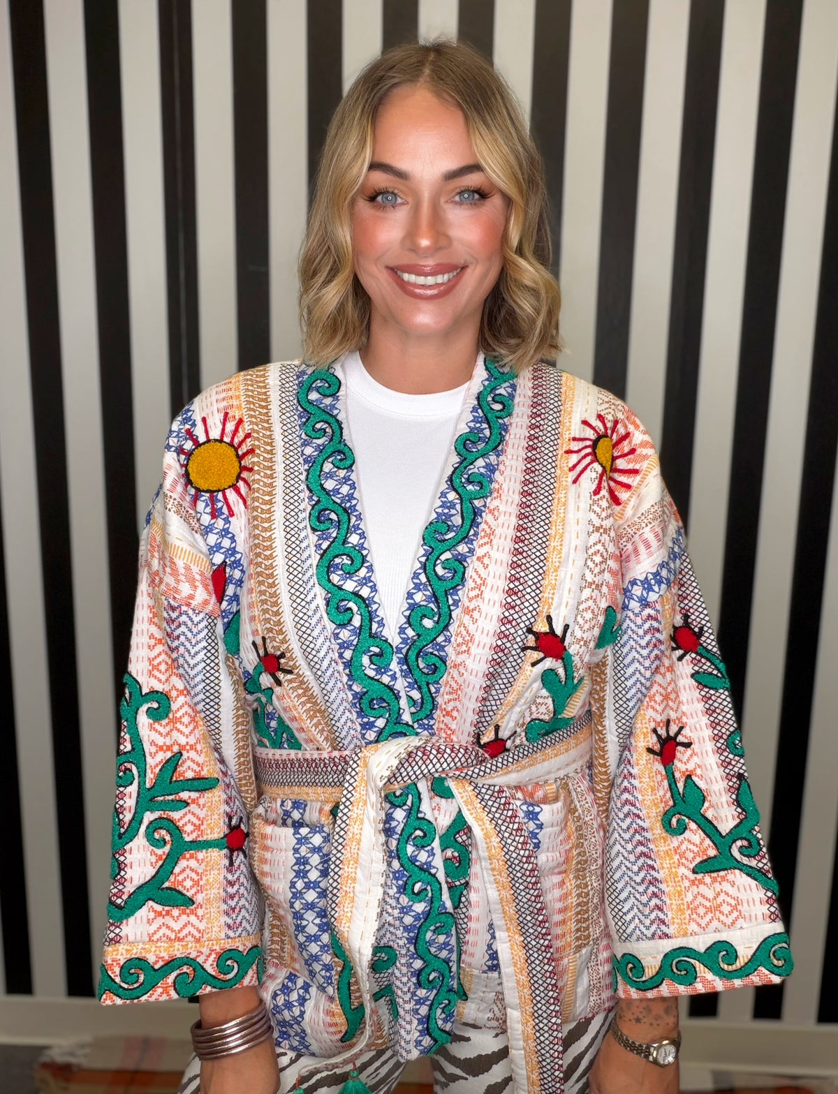 A smiling blonde woman wearing a colorful patterned kimono-style robe with embroidered sun motifs and green vine details over a white t-shirt, posed against a black and white striped background.