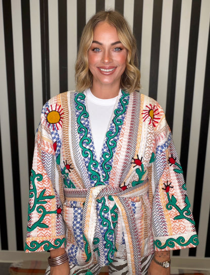 A smiling blonde woman wearing a colorful patterned kimono-style robe with embroidered sun motifs and green vine details over a white t-shirt, posed against a black and white striped background.