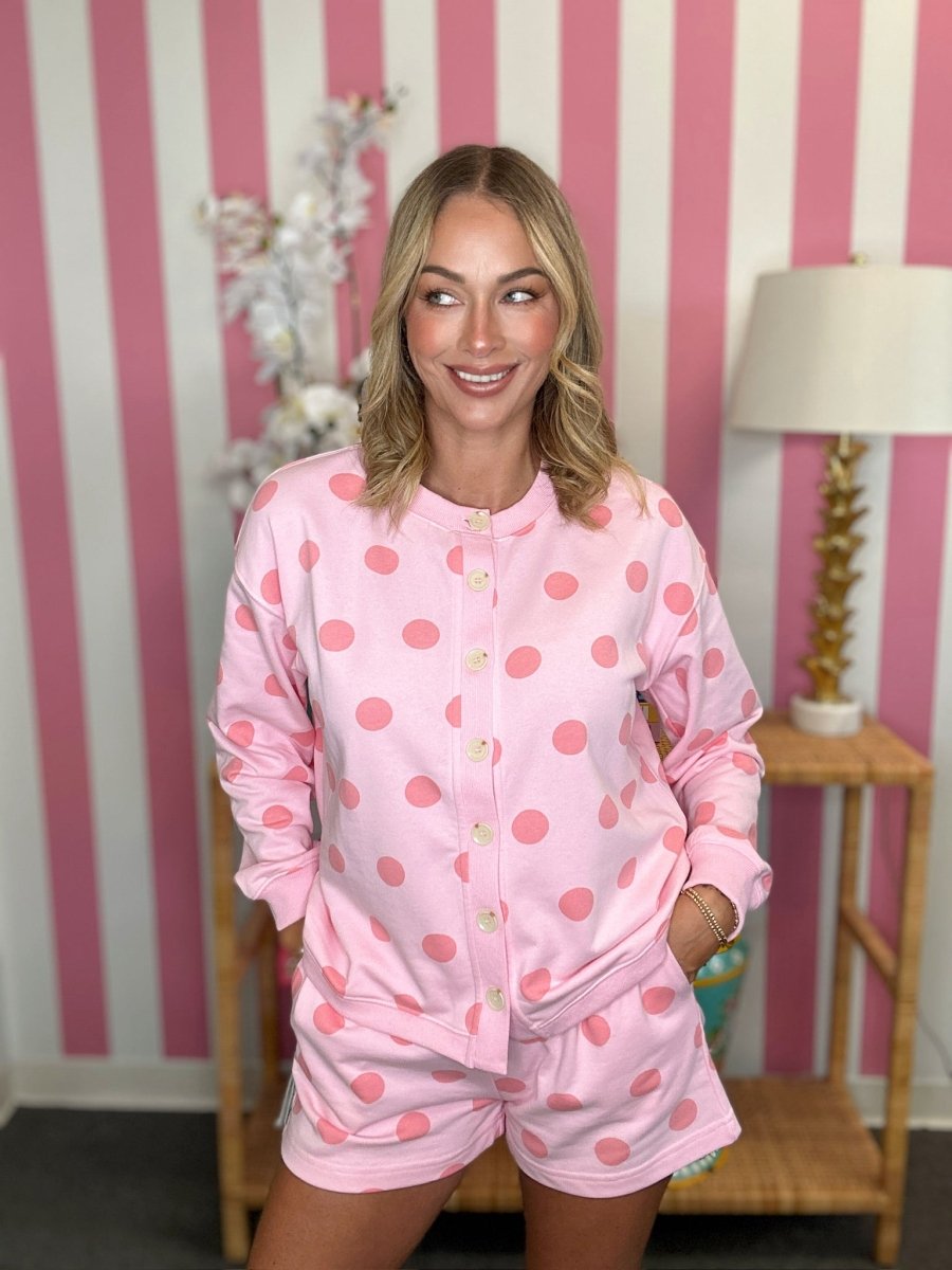 A woman modeling a pink two-piece pajama set with large pink polka dots, consisting of a long-sleeve button-front top and matching shorts, set against a pink and white striped wall.