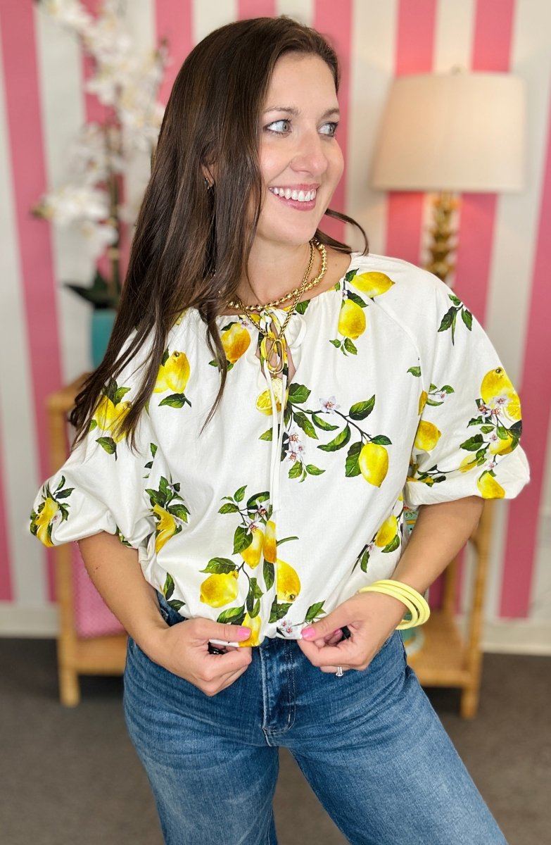 Woman wearing a white blouse with yellow lemon print and blue jeans in a room with striped wallpaper.