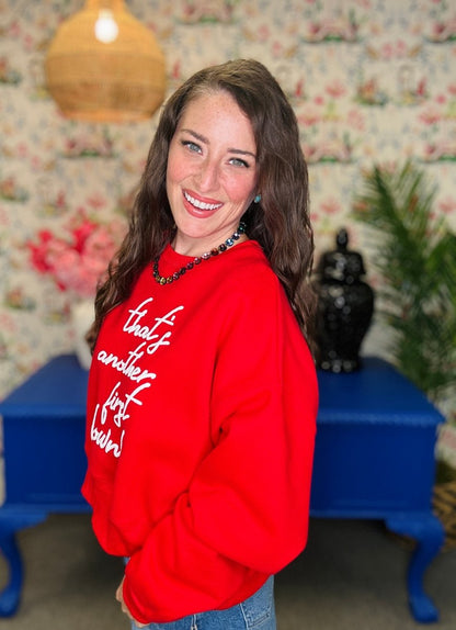 A woman smiling and looking over her shoulder while wearing a bright red sweatshirt with white cursive text that reads 'that's another day of hell!' in a decorated room with floral wallpaper, a blue table, and a plant.