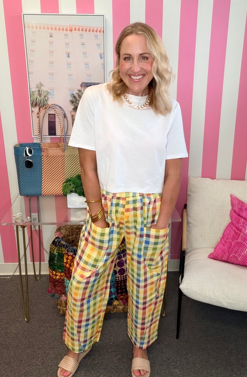 A woman stands in a room with pink and white striped walls, wearing a white short-sleeve t-shirt and colorful plaid cropped pants. She is accessorized with a gold chain necklace and bracelets, standing next to a small table with a blue bag and a white chair with a pink pillow.