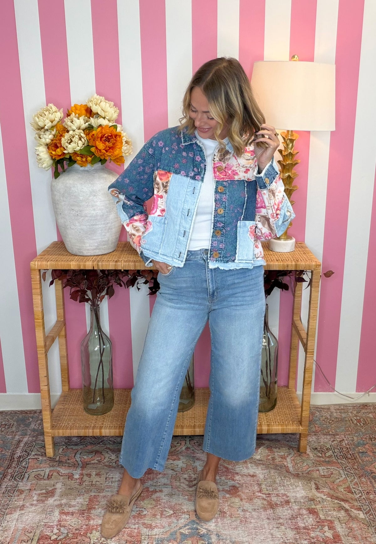 A woman stands in a room with pink and white striped wallpaper, wearing a colorful patchwork denim jacket, light blue cropped jeans, and tan loafers. A wooden side table with flowers and a lamp is behind her.