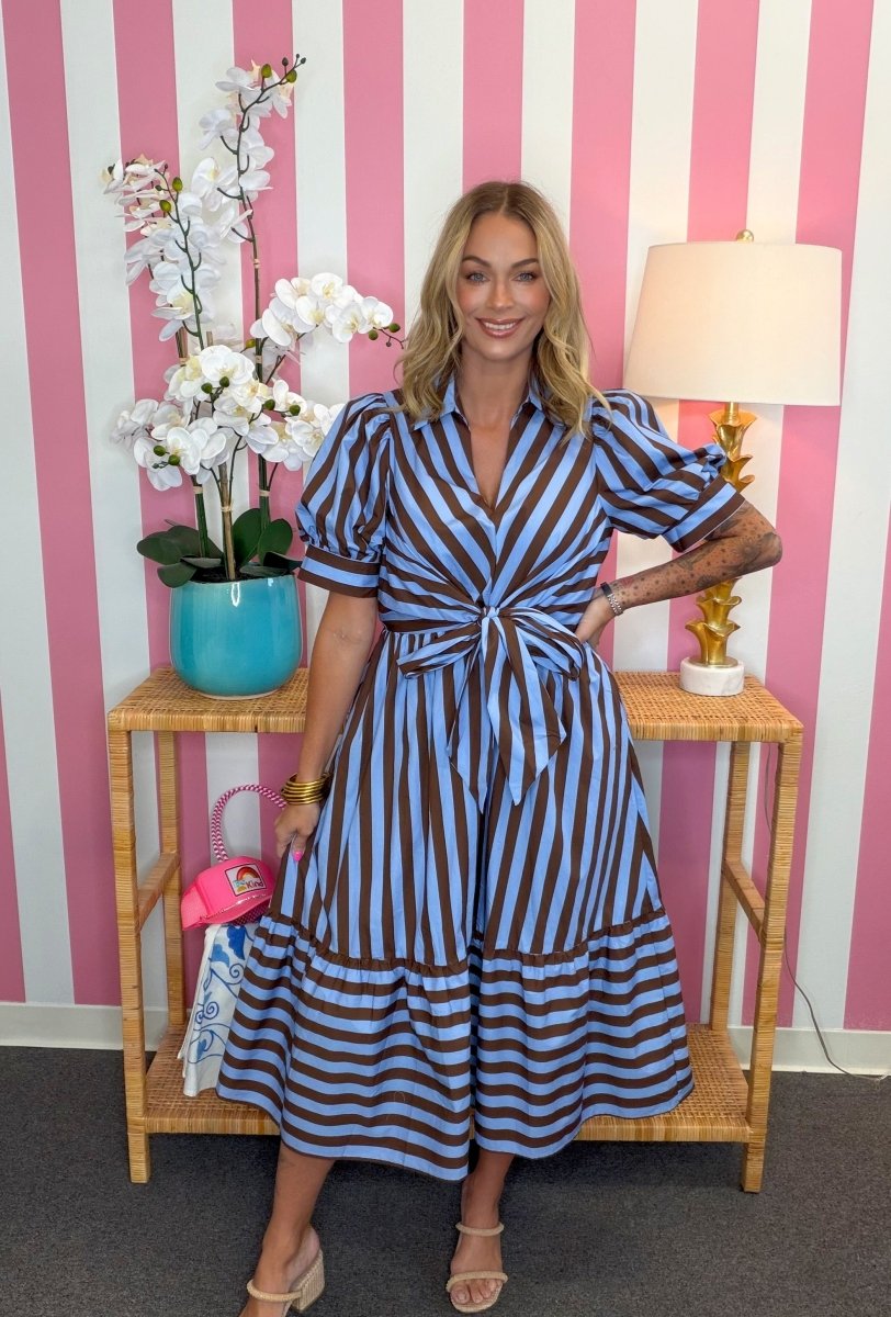 A woman models a blue and brown striped midi dress with puff sleeves and a tie waist, standing in front of a pink and white striped wall next to a side table with a lamp and flowers.