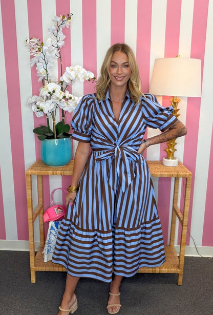 A woman models a blue and brown striped midi dress with puff sleeves and a tie waist, standing in front of a pink and white striped wall next to a side table with a lamp and flowers.