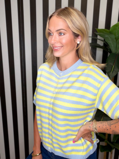 A woman wearing a yellow and light blue striped short-sleeve knit cardigan sweater, standing in front of a black and white striped wall with a green plant in the background.