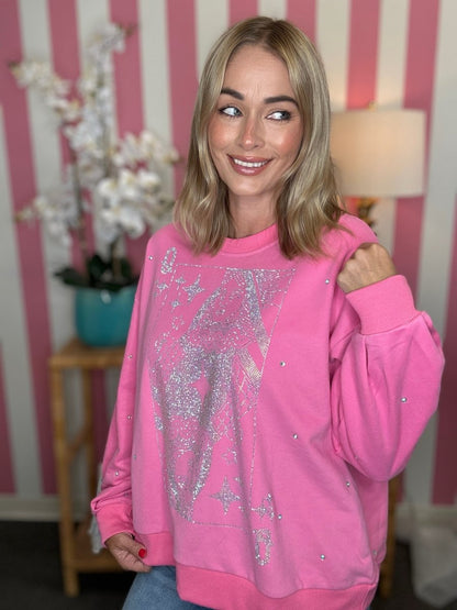 A woman wearing a bright pink crewneck sweatshirt with a large silver glittery graphic on the front and small rhinestone embellishments, posed in front of a pink and white striped wall.