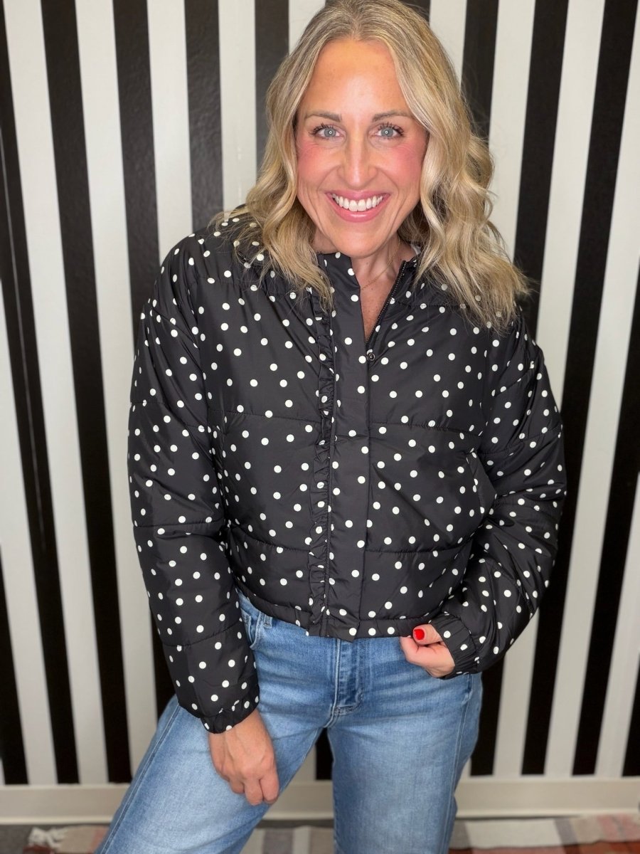 A smiling blonde woman wearing a black puffer jacket with white polka dots and blue jeans, standing in front of a black and white vertically striped wall.