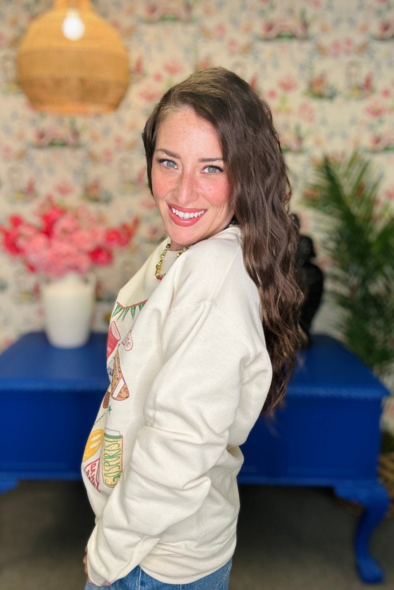 A woman with long brown hair smiling and looking back over her shoulder, wearing a cream-colored graphic sweatshirt and blue jeans in a decorated room with floral wallpaper, a blue bench, and a hanging lamp.