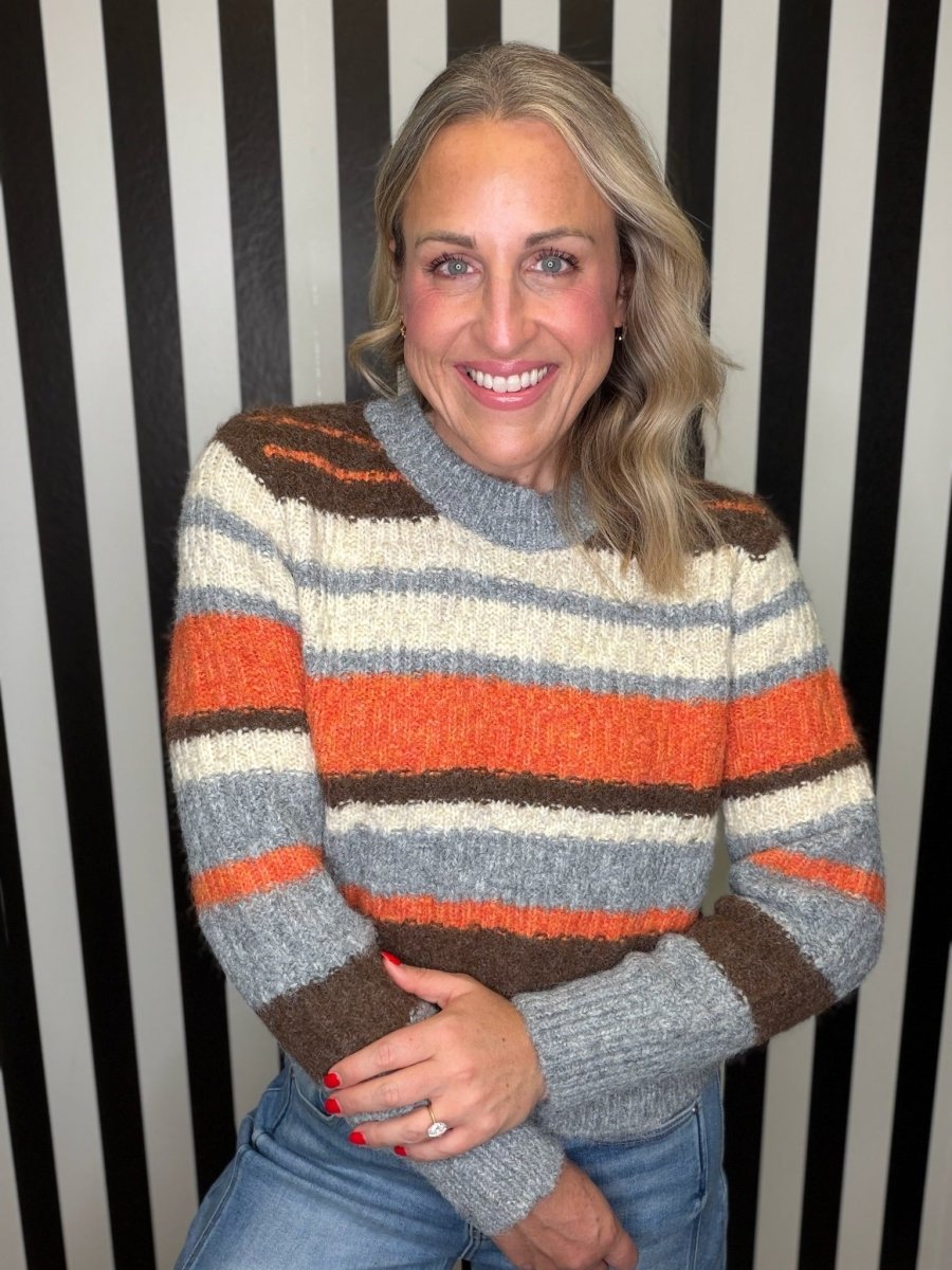 A smiling woman with blonde hair wearing a fuzzy, striped knit sweater in orange, brown, gray, and cream, paired with blue jeans, standing in front of a black and white striped wall.