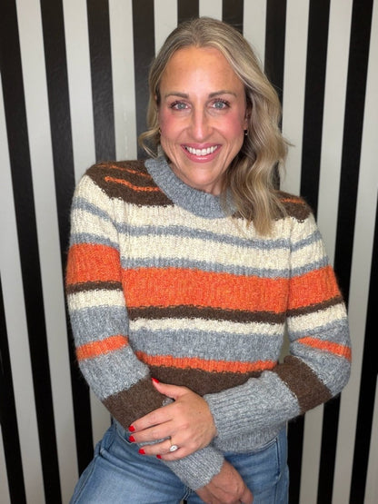 A smiling woman with blonde hair wearing a fuzzy, striped knit sweater in orange, brown, gray, and cream, paired with blue jeans, standing in front of a black and white striped wall.