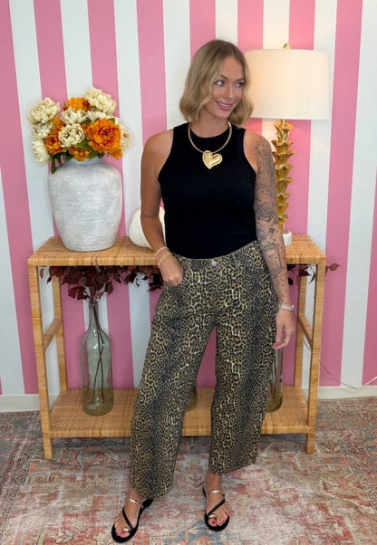 A woman stands in a room with pink and white striped wallpaper, wearing a black sleeveless top and wide-leg cropped pants with a brown and black leopard print. She is posing in front of a wooden console table decorated with a vase of flowers and a lamp.
