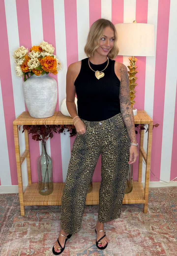 A woman stands in a room with pink and white striped wallpaper, wearing a black sleeveless top and wide-leg cropped pants with a brown and black leopard print. She is posing in front of a wooden console table decorated with a vase of flowers and a lamp.