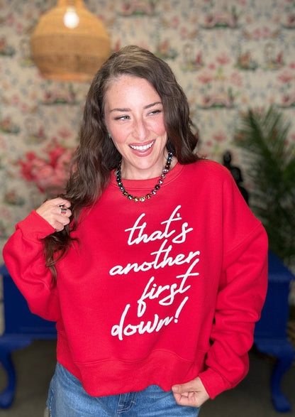A woman smiling while wearing a red crewneck sweatshirt with the phrase 'that's another first down!' printed in white script across the front, paired with blue jeans in a decorated indoor setting.