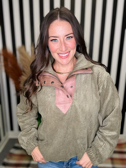 A woman wearing a ribbed olive green pullover sweater with a dusty pink mock neck and button placket, paired with blue jeans, standing in front of a black and white striped background.