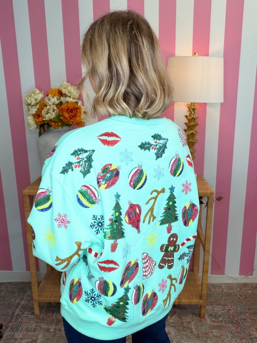 A woman viewed from behind wearing a light aqua Christmas sweatshirt with colorful holiday prints, standing in front of pink and white striped wallpaper.