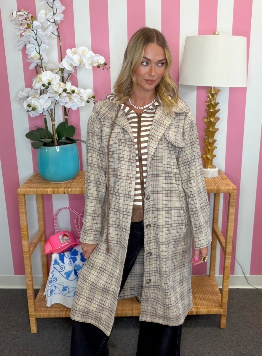 A woman models a long beige and brown plaid coat over a striped top, standing in front of a pink and white striped wall with a lamp and side table in the background.