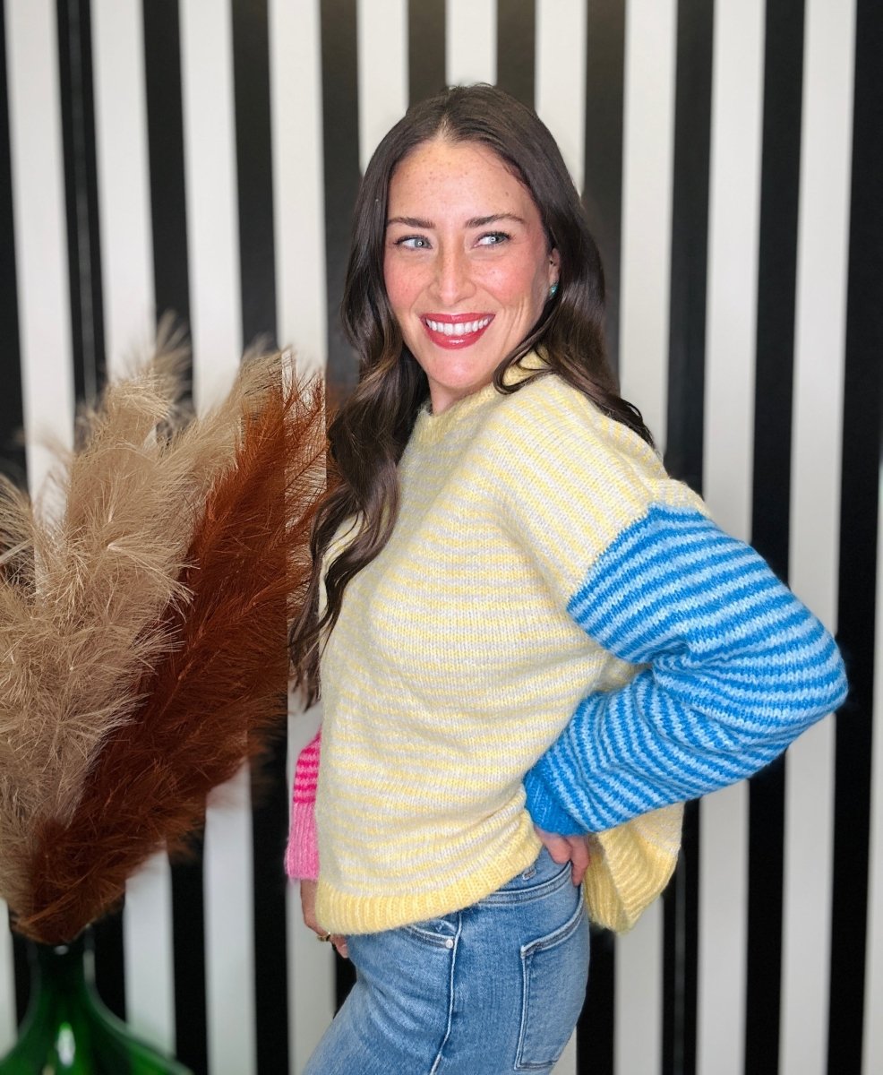 A woman modeling a pastel knit sweater with a pale yellow body and bright blue striped sleeves, paired with blue jeans, standing in front of a black and white striped wall.
