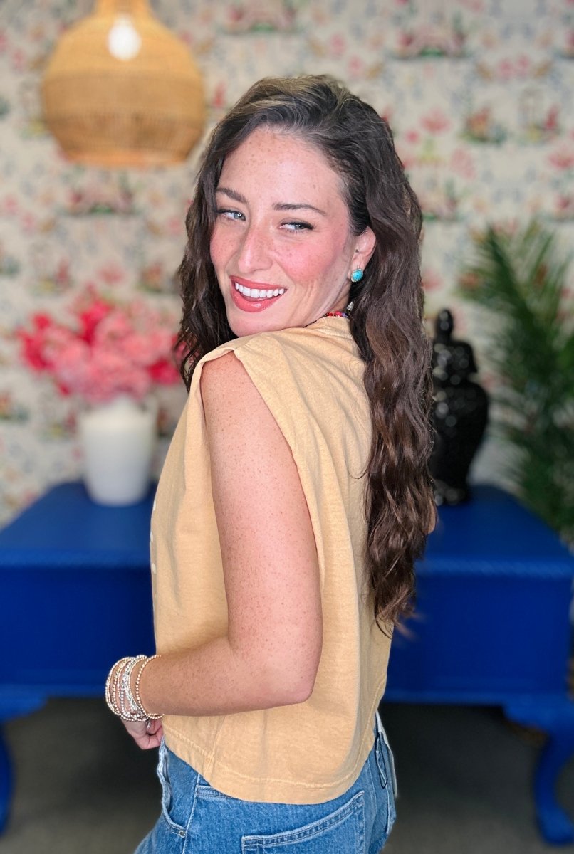A woman with long wavy brown hair smiles while looking over her shoulder, wearing a sleeveless mustard yellow top and blue jeans in a decorated room with floral wallpaper and a blue table.