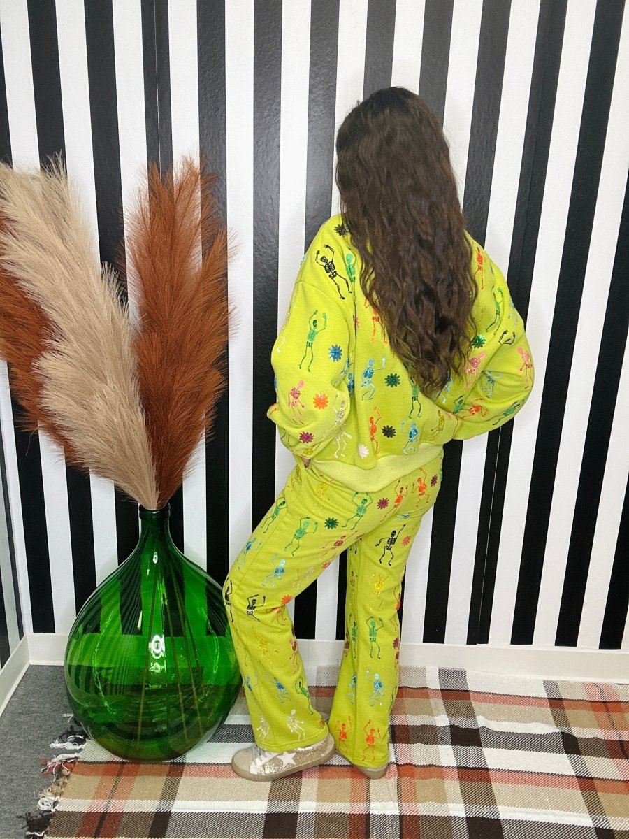 A person wearing a bright yellow-green pajama set with colorful floral and abstract prints, standing in front of a black and white striped wall next to a large green vase with pampas grass.