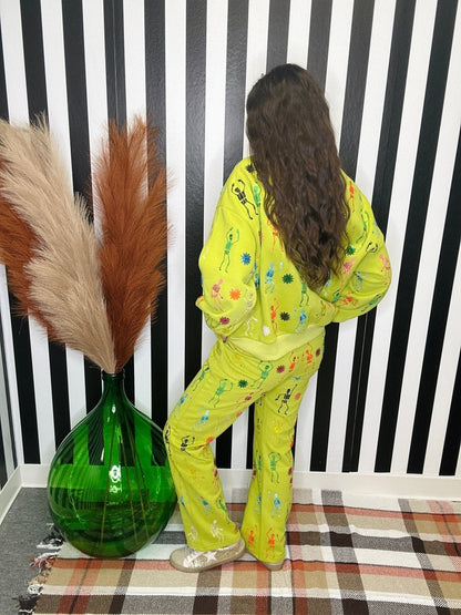 A person wearing a bright yellow-green pajama set with colorful floral and abstract prints, standing in front of a black and white striped wall next to a large green vase with pampas grass.