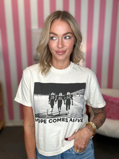 A woman wearing a cream-colored graphic t-shirt with a black and white football player print and the text 'HOPE COMES ALIVE', paired with blue jeans, standing in front of pink and white striped wallpaper.