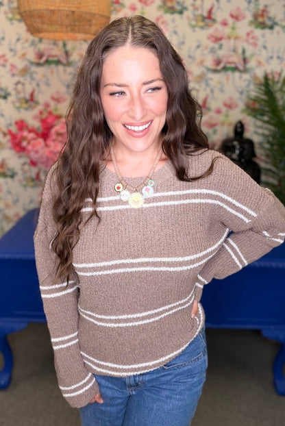 A woman with long wavy brown hair smiling while wearing a brown knit sweater with white horizontal stripes and blue jeans, standing in front of a floral wallpaper background.