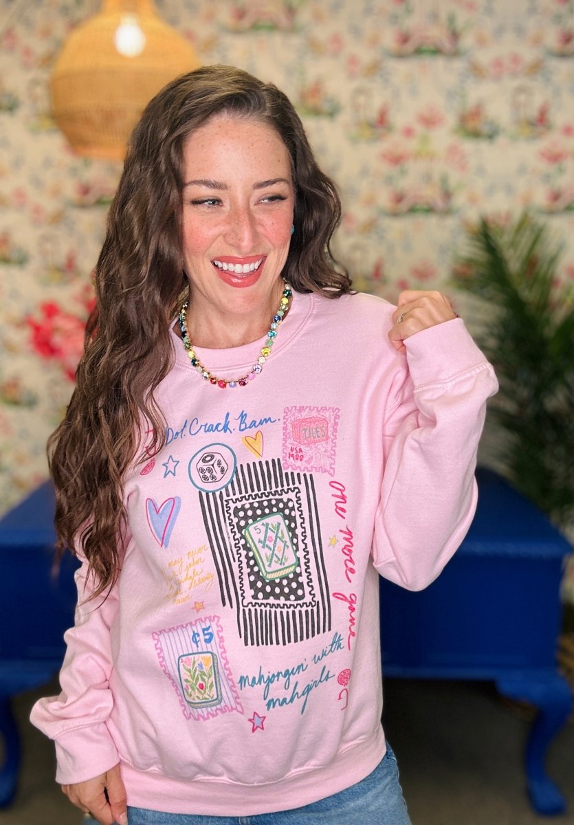 A woman smiling and wearing a light pink crewneck sweatshirt with a colorful graphic patch design, set against a vintage floral wallpaper background.