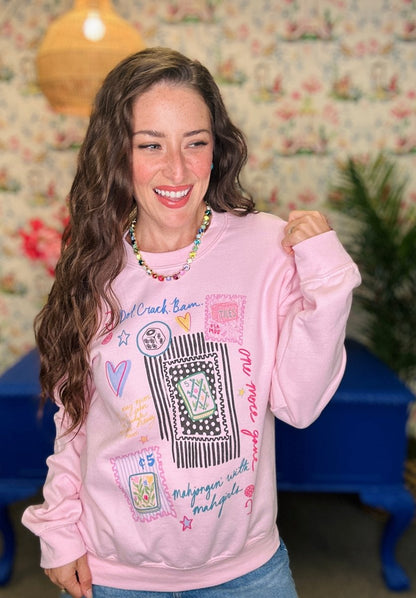 A woman smiling and wearing a light pink crewneck sweatshirt with a colorful graphic patch design, set against a vintage floral wallpaper background.