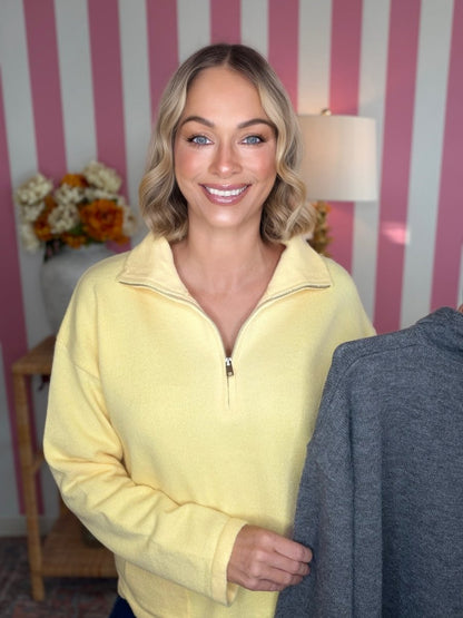 A smiling blonde woman wearing a pale yellow quarter-zip fleece pullover stands indoors against pink-and-white striped wallpaper, holding a dark gray pullover in front of the camera.