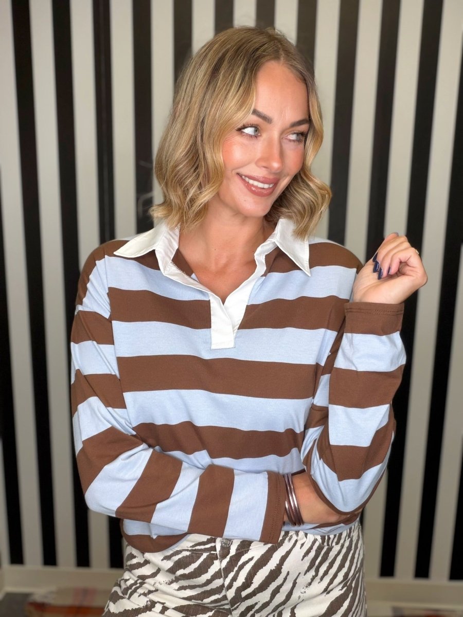 A woman wearing a brown and light blue horizontally striped long-sleeve rugby shirt with a white collar, paired with brown and white zebra-print trousers, posed against a black and white striped background.