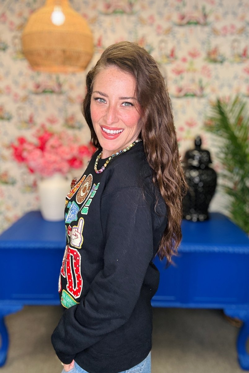 A woman with long brown hair smiles while looking over her shoulder, wearing a black crewneck sweatshirt with colorful graphic patches on the front. She is standing in a room with floral wallpaper, a blue table, and a hanging lamp.