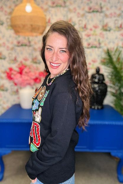 A woman with long brown hair smiles while looking over her shoulder, wearing a black crewneck sweatshirt with colorful graphic patches on the front. She is standing in a room with floral wallpaper, a blue table, and a hanging lamp.