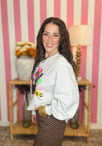 A woman smiling and posing in a light gray sweatshirt with a colorful graphic print on the front, paired with brown and white polka dot pants, set against a pink and white striped wall background.