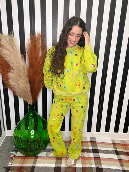 A woman modeling a bright yellow-green matching lounge set with colorful floral prints, standing in front of a black and white striped wall next to a green glass vase with pampas grass.