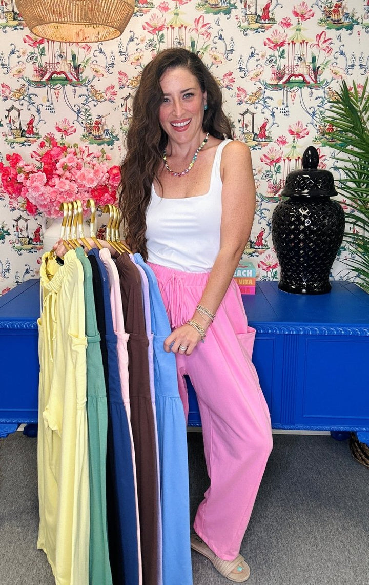 A woman stands indoors holding a rack of colorful sleeveless tops. She is wearing a white tank top and bright pink wide-leg pants.